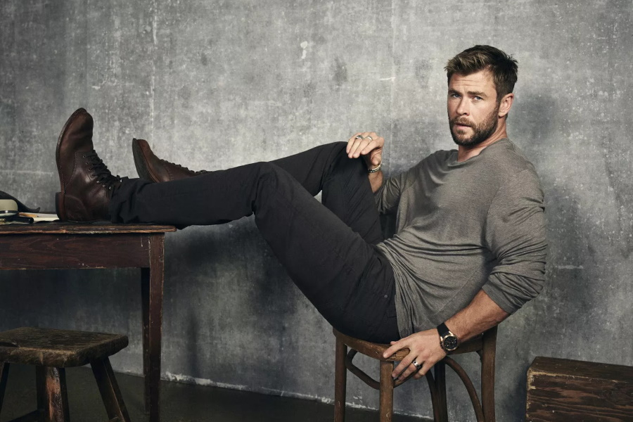 Chris Hemsworth sitting on a wooden chair with his feet propped up on a wooden desk