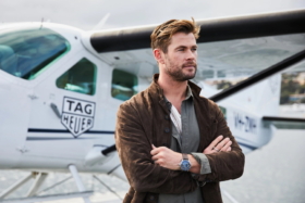 Medium shot Chris Hemsworth with his arms crossed and a plane behind him in the background