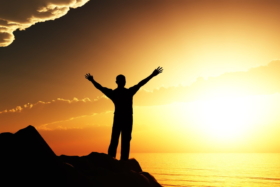 Silhouette of a man on a hill with his arms raised in air in front of sun