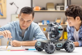 A man using a compass on table as a boy watches him. A DJI Robomaster robot on table