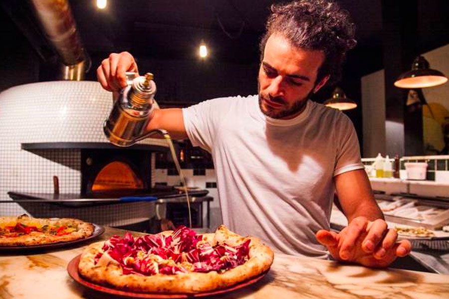 A man pouring a liquid out of a metal kettle on a pizza on table