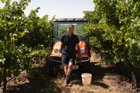 Jock Harvey in middle of vineyard leaning his back on the front hood of a small farm car