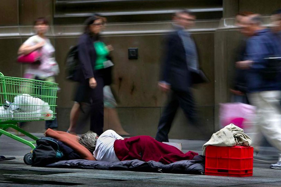 A homeless person sleeping on pavement as people walk by