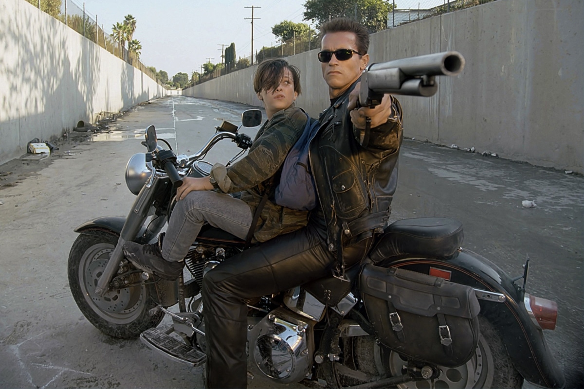 Motorcycle rider in leather jacket aims gun, with passenger holding on, in a concrete-lined urban setting.