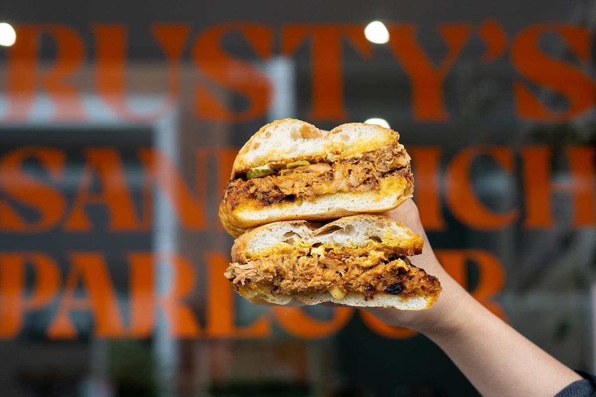 hand holding sandwich in front of rustys sandwich parlour 