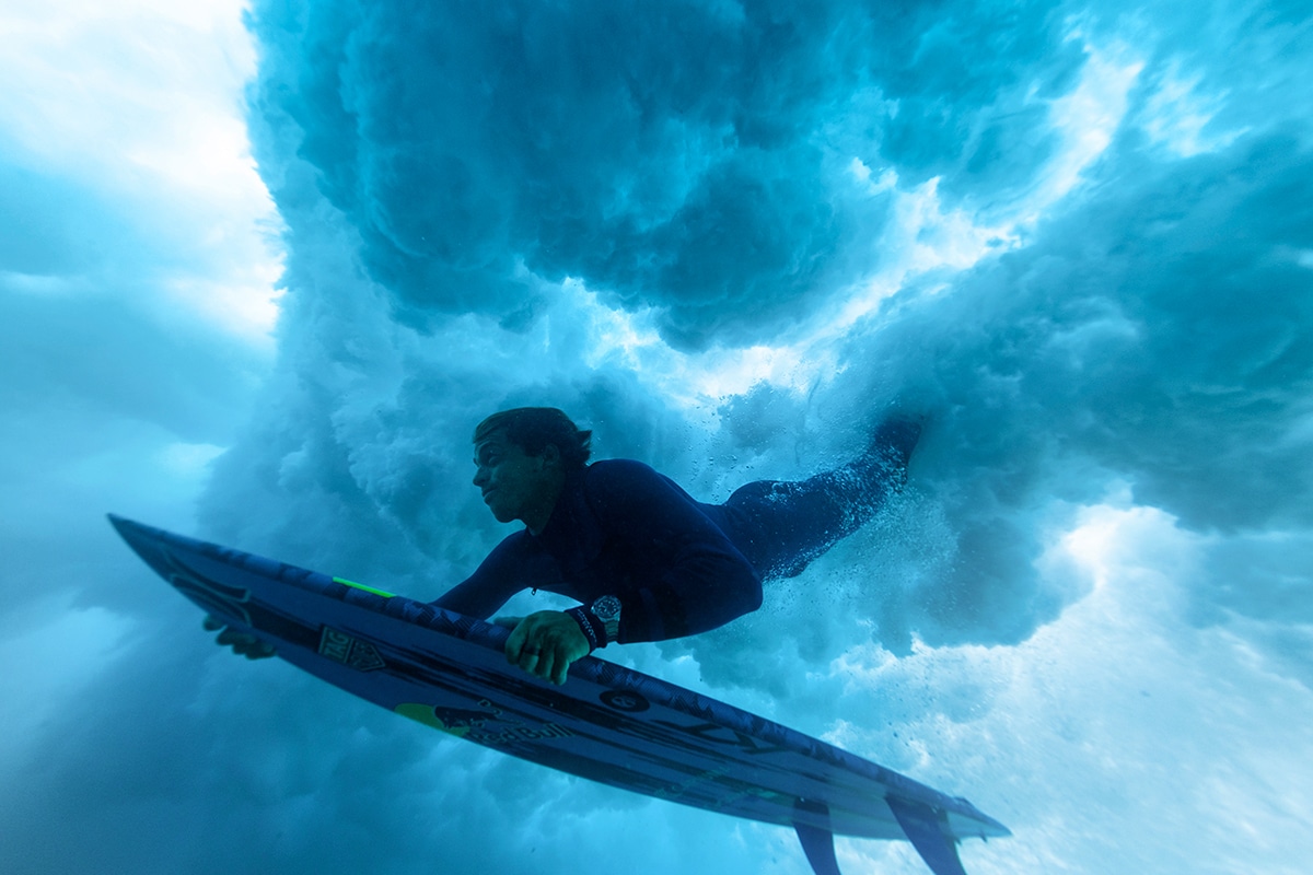 Kai Lenny underwater surfing with a blue surfboard, wearing a wetsuit and a Tag Heuer watch, surrounded by swirling ocean waves.