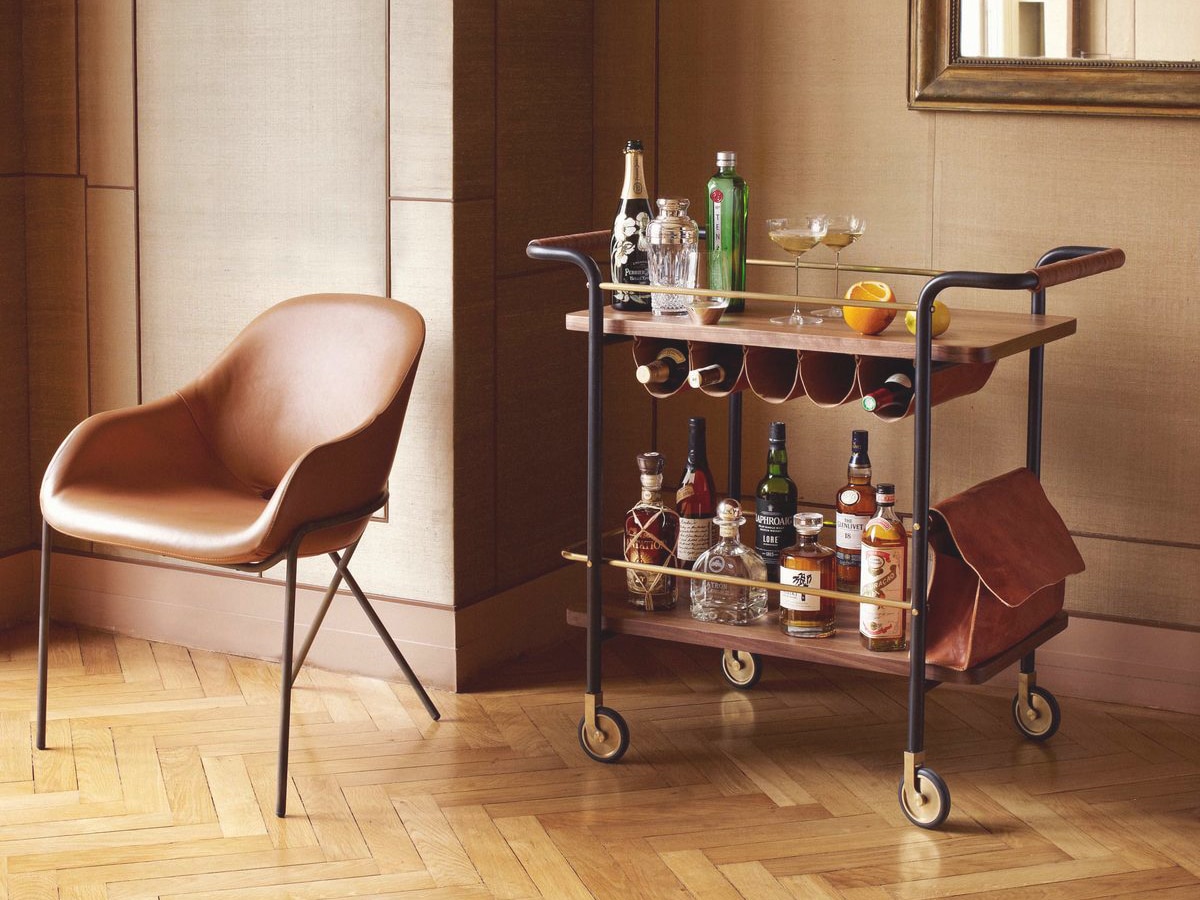 Valet bar cart with liquor bottles, glasses, and an orange, beside a brown leather chair on wooden flooring.