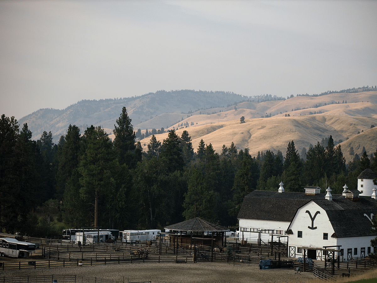 Ranch landscape with white barn, surrounded by trees and rolling hills under a cloudy sky.