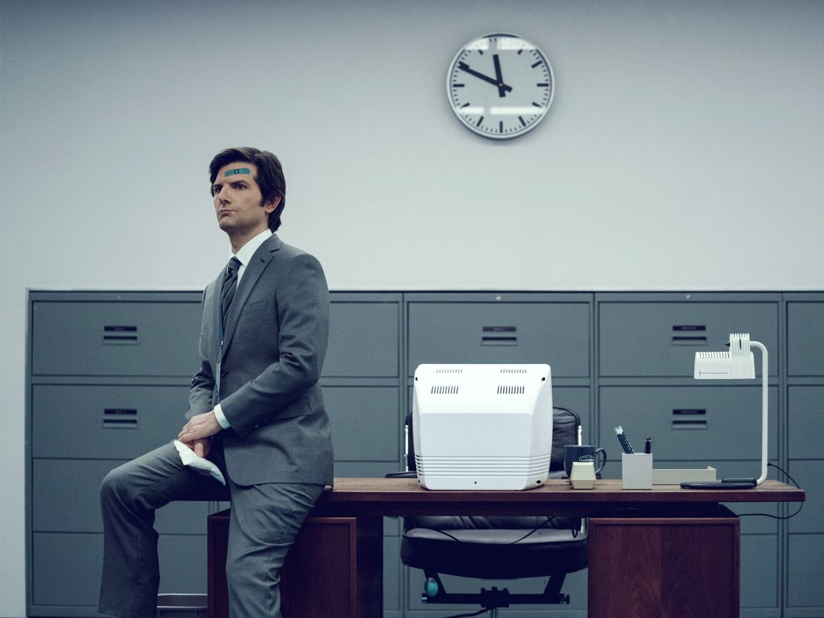 Adam Scott in a suit sits on an office desk with a clock on the wall and filing cabinets in the background.
