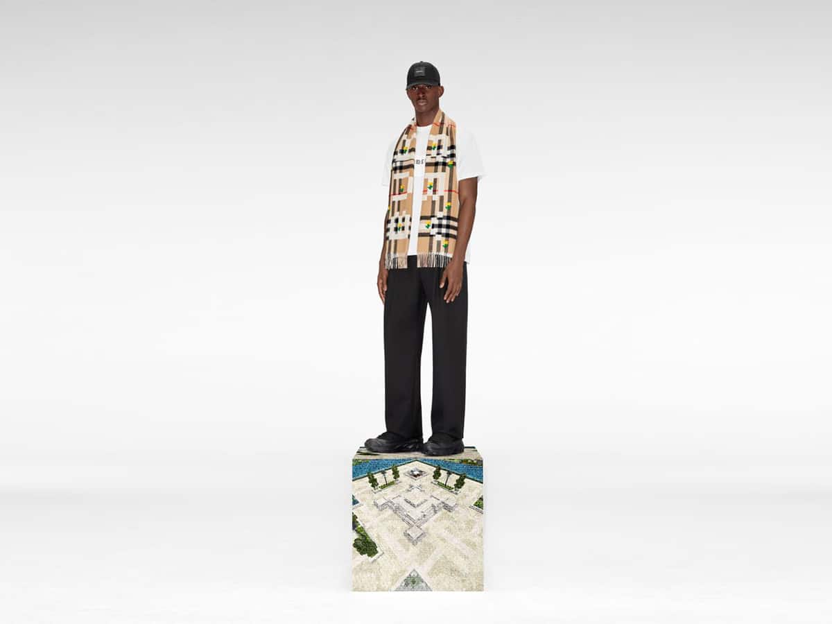 A person wearing a Burberry scarf and black cap stands on a Minecraft-themed block in a white studio.