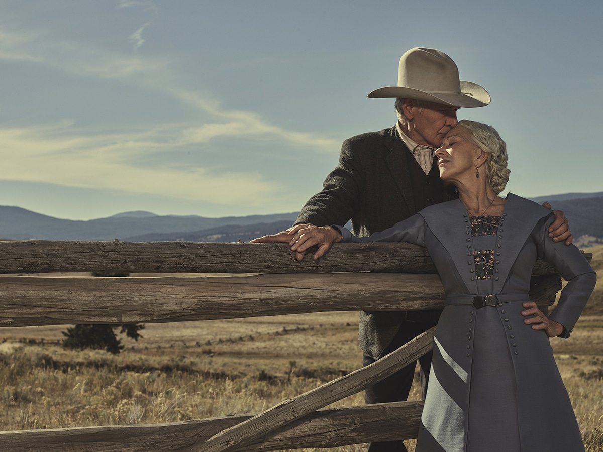 Elderly couple in period clothing embrace by a wooden fence with a scenic mountain backdrop.