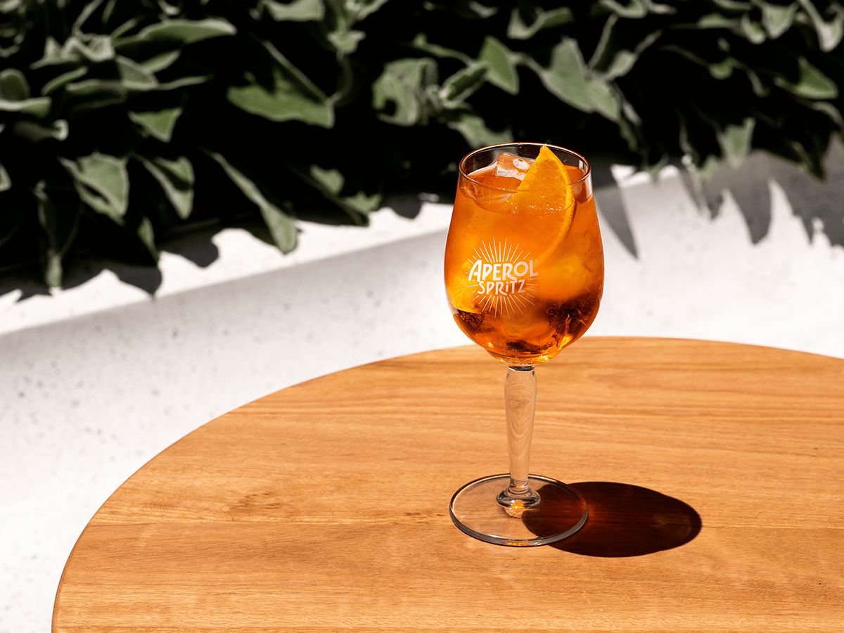 Aperol Spritz glass with orange slice on wooden table, set against lush green foliage in bright sunlight.