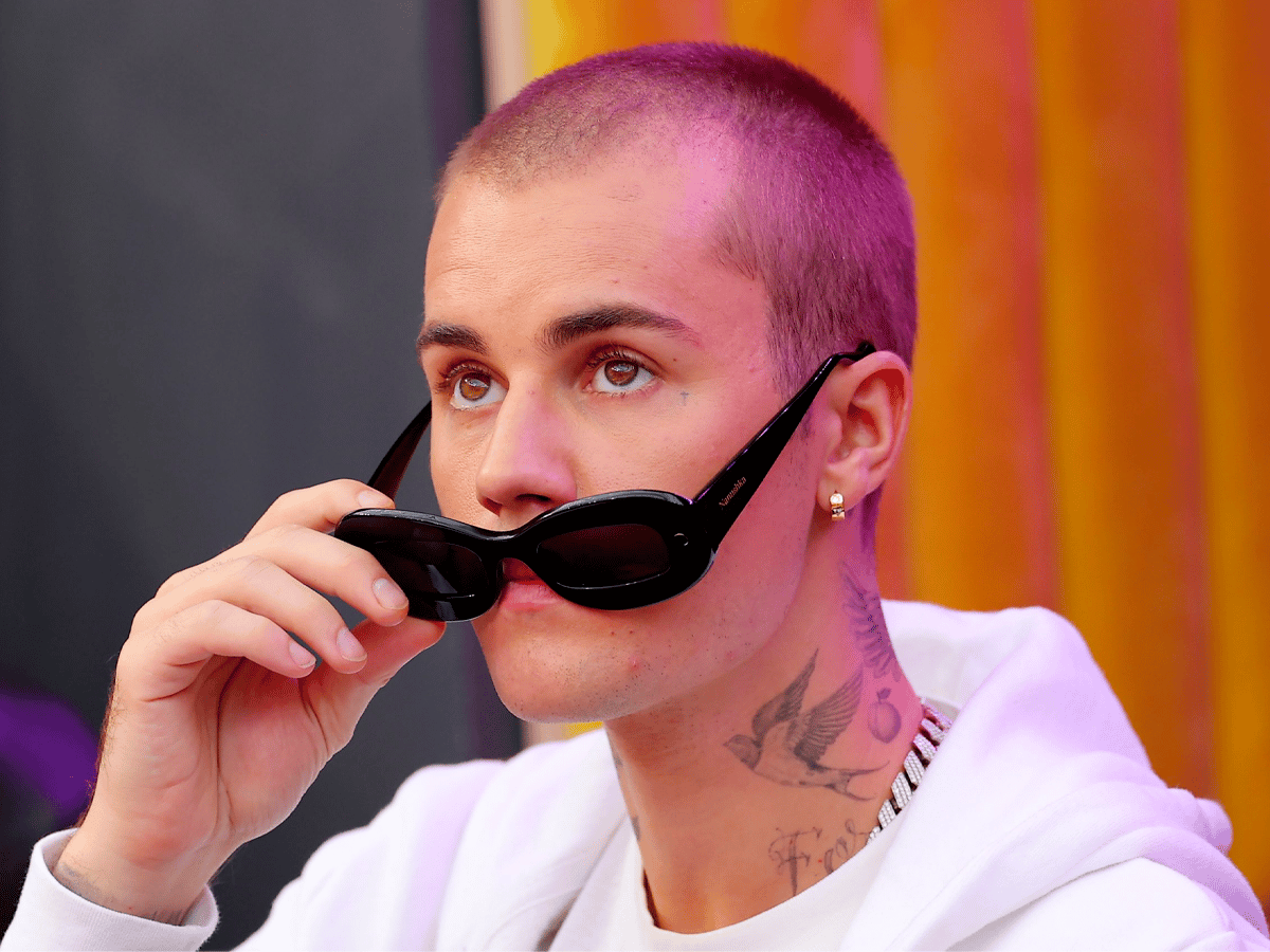 A person with a buzz cut lifts black sunglasses, wearing a white hoodie and gold chain, with a bird tattoo on their neck.