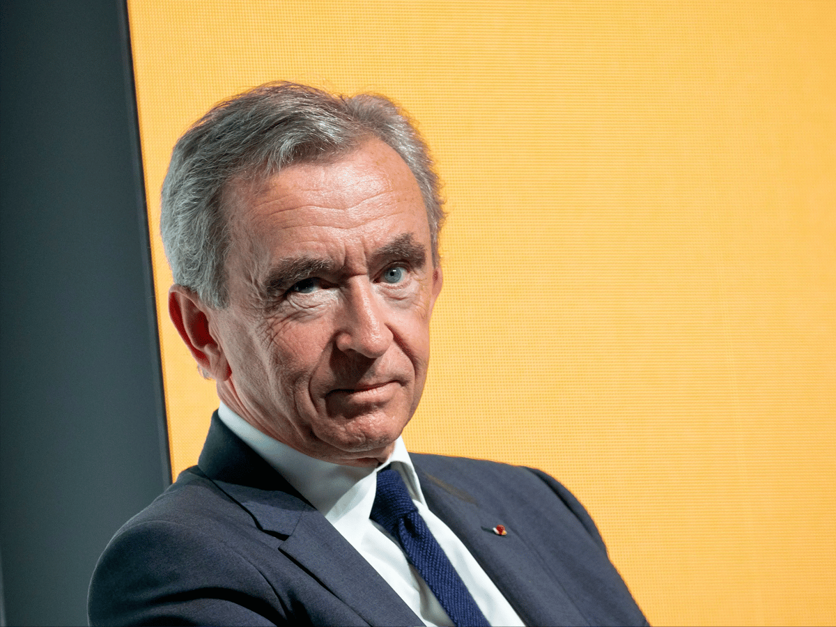 A man in a dark suit and tie sits against a bright orange background, looking thoughtfully to the side.