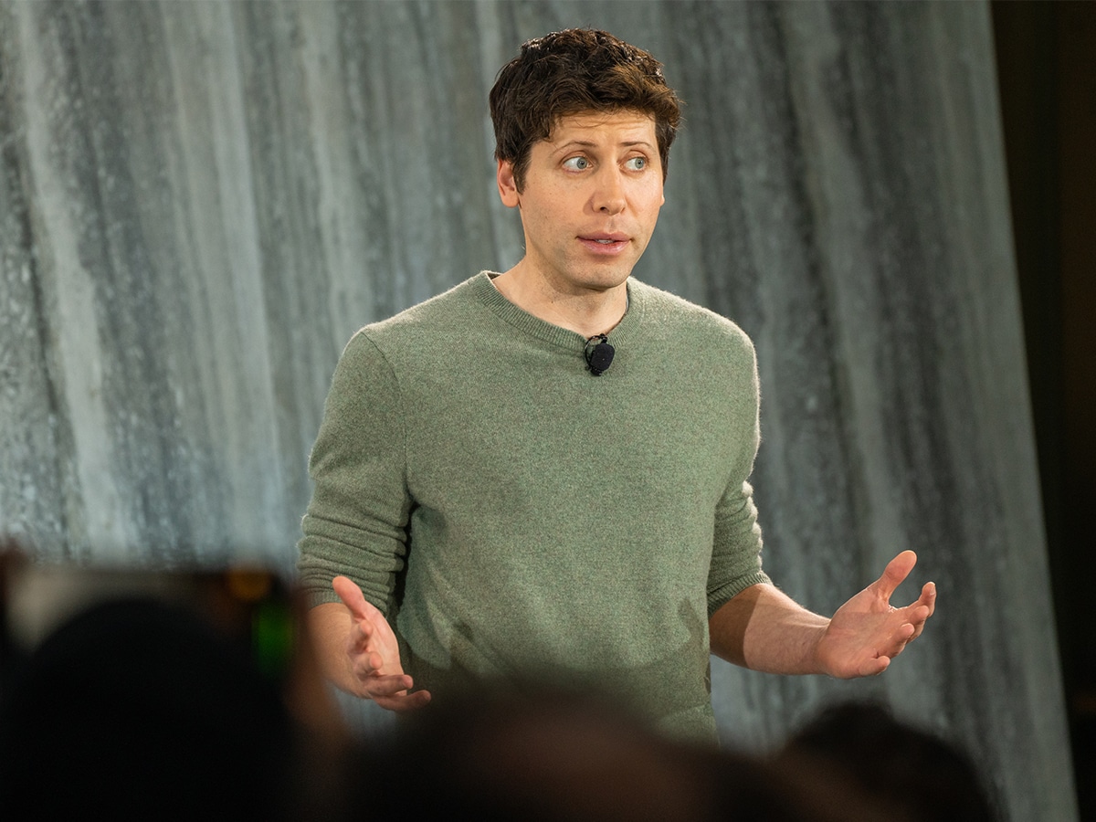 A man in a grey sweater gestures with both hands, standing in front of a textured grey wall.