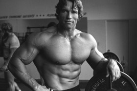 Shirtless Arnold Schwarzenegger with one hand on his hip and the other resting on a barbell