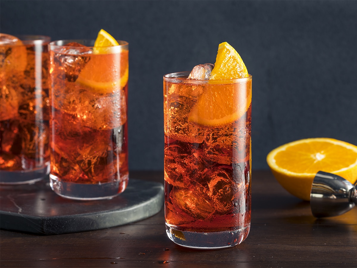 Three glasses of Americano cocktail on the rocks with orange garnish set on a dark wooden bar with a dark background