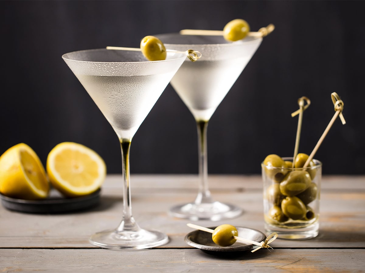 Two glasses of dry martini cocktail served with an olive set on a grey wooden bar with dark background