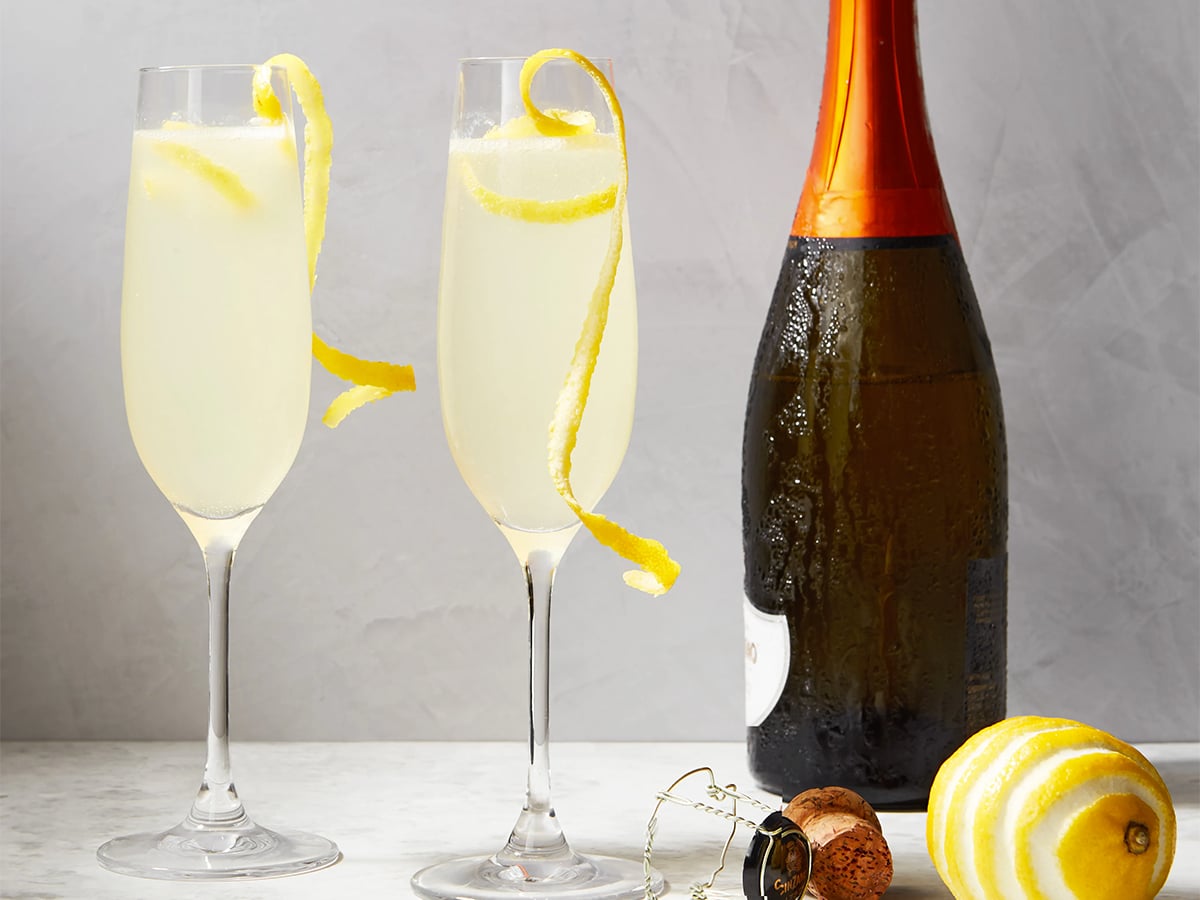 Two glasses of French 75 served with a lemon twist garnish beside a bottle set on a matte marble background