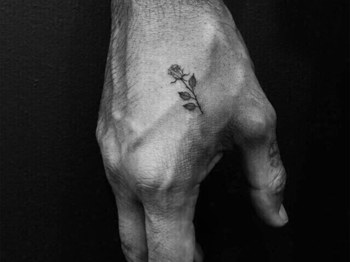 Small black and gray tattoo of rose with thorny stem and three petals on a man's hand