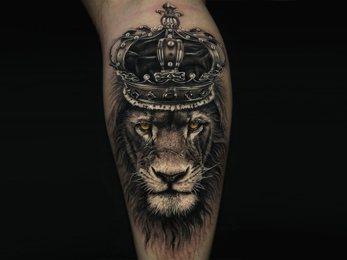 Black and gray tattoo of a lion with yellow eyes wearing a crown on forearm