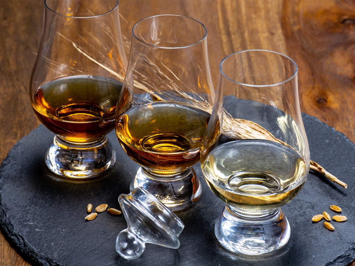 Three glasses of whiskey varying in color set on a dark platter on a wooden table