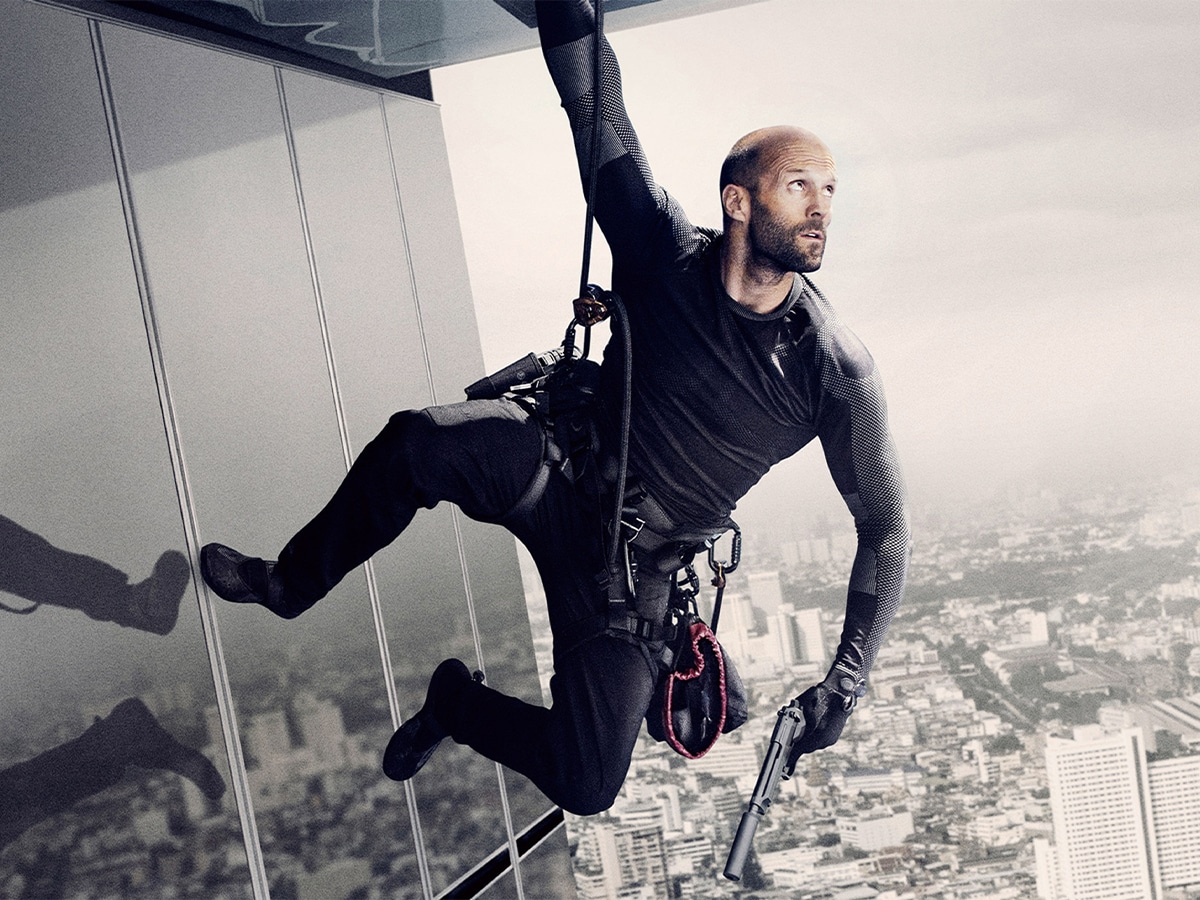 Jason Statham hanging from a building grabbing a safety rope with one hand and holding a gun