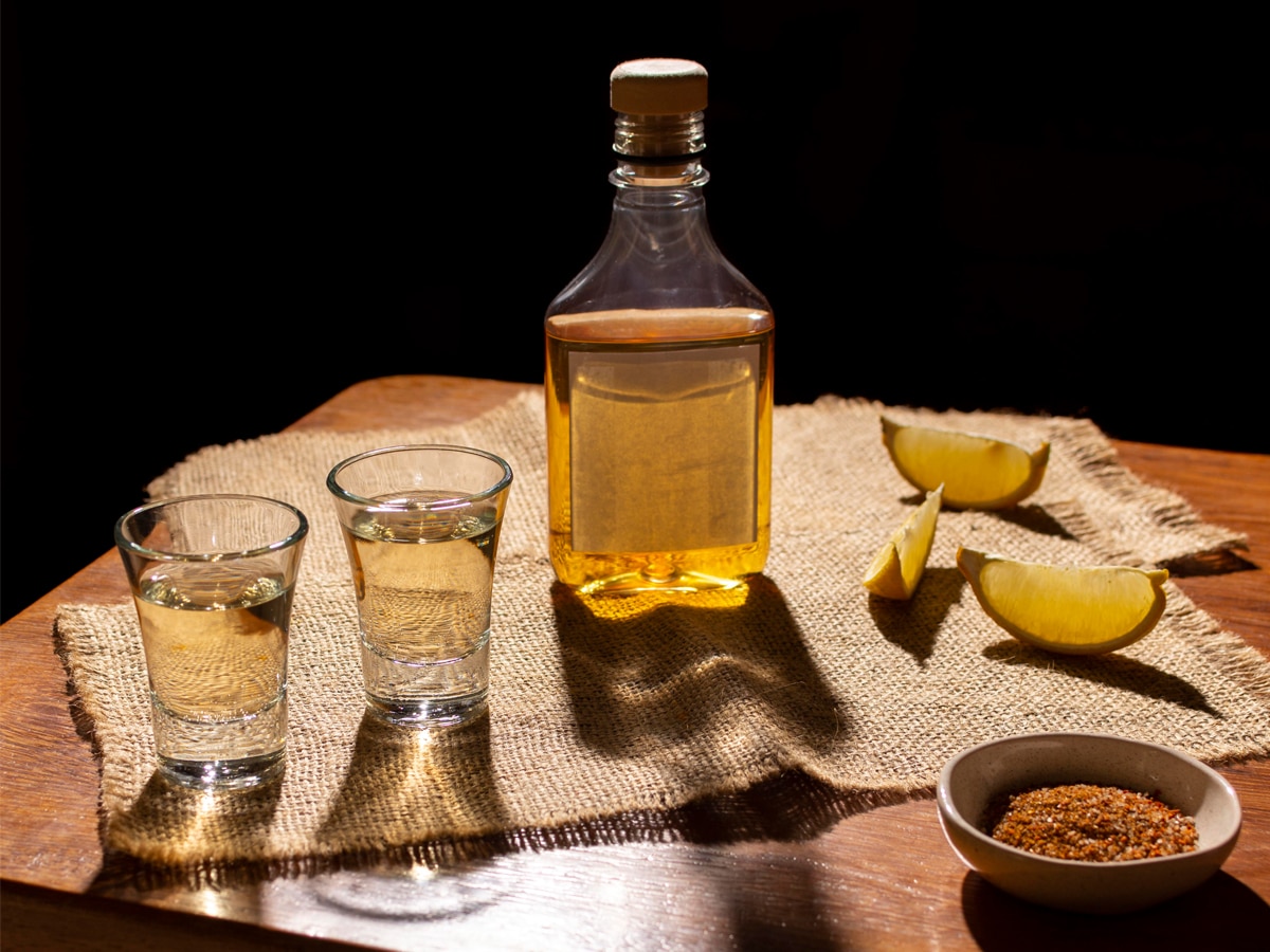 A clear bottle and two crystal glasses filled with mezcal with three lemon slices and a dip bowl of Sal De Gusano on a wooden table
