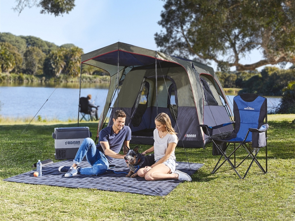 Couple sitting on a picnic blanket with their dog and ALDI's 4 Person Pop-Up Tent set up behind them