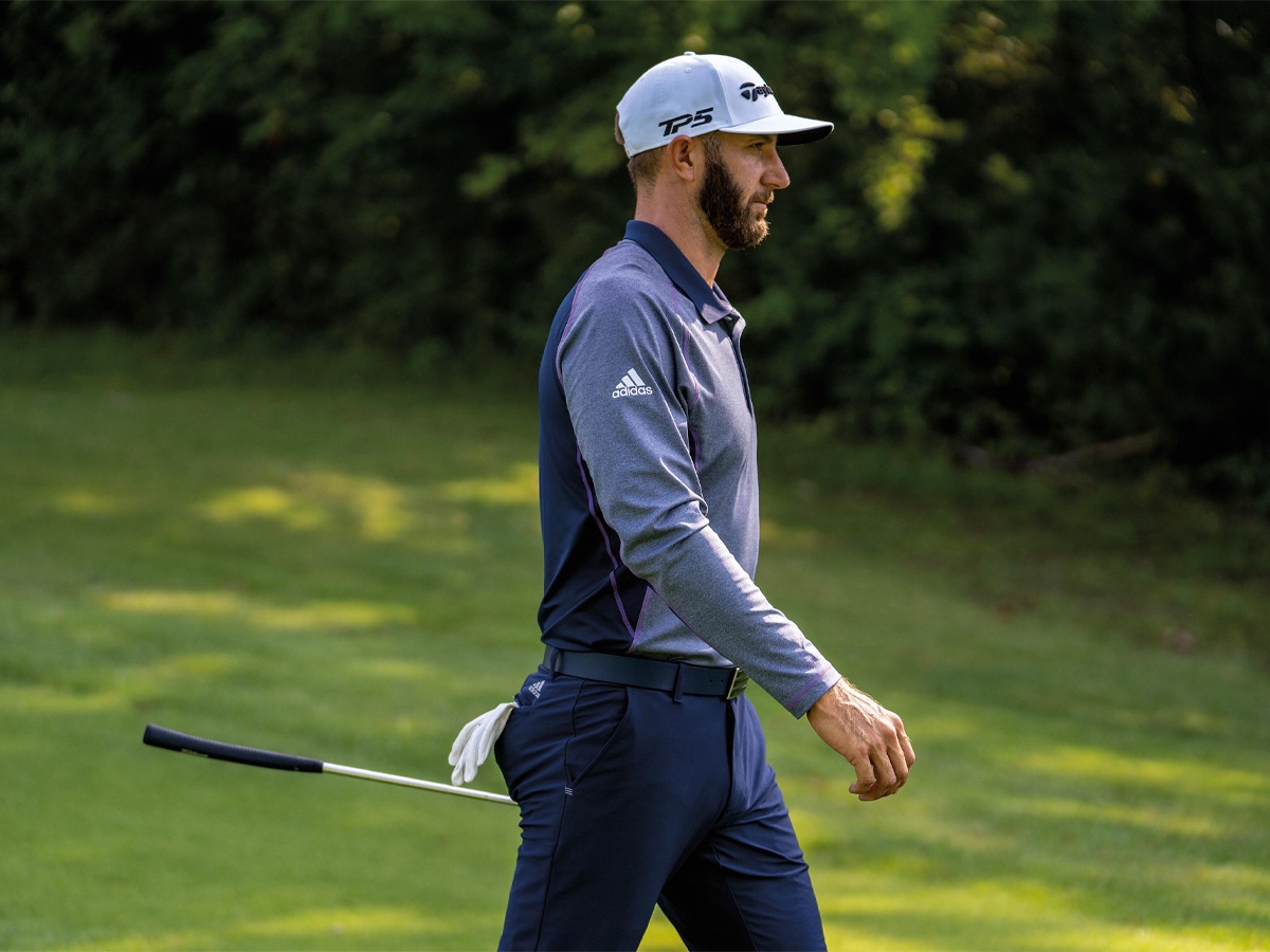 A golfer in a dark blue and violet long sleeve shirt, white cap, and dark blue pants