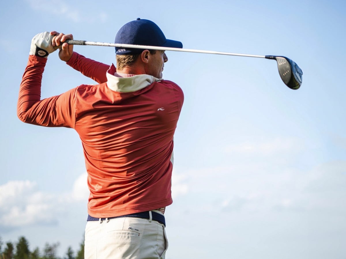 A golfer in a red jacket, white pants, blue cap, and a white golf glove