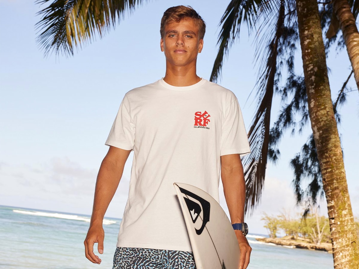 Male model holding a surfboard and wearing a white logo shirt and printed shorts