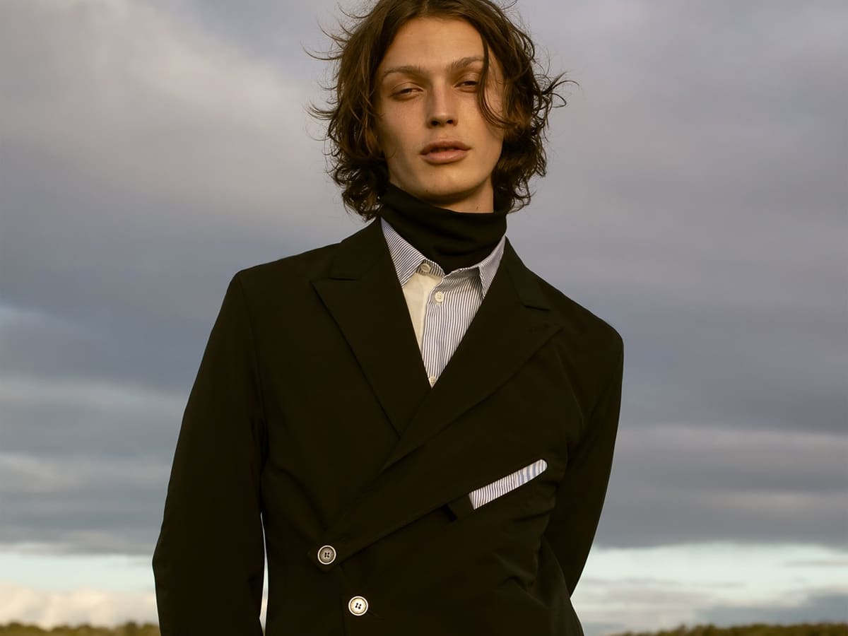 Male model in a black turtleneck underneath blue and white buttoned shirt underneath black coat
