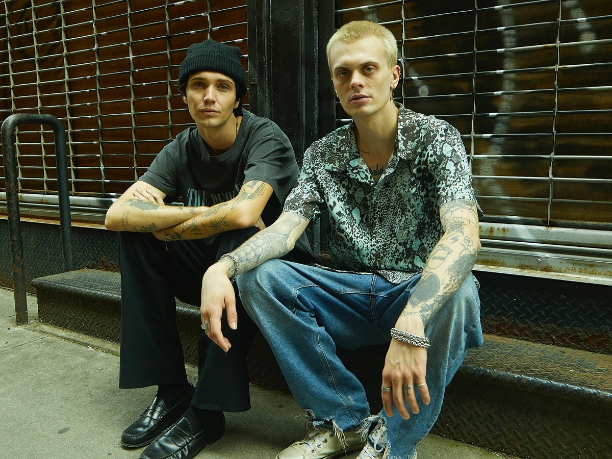Male model in a printed shirt and denim pants beside another male model in a black beanie hat, black graphic t-shirt, and black pants