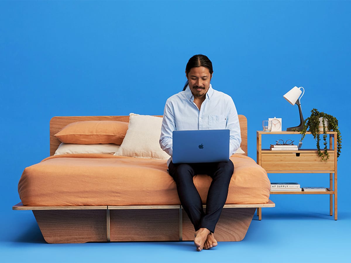 Man with laptop on his lap sitting on the edge of his bed with blue background