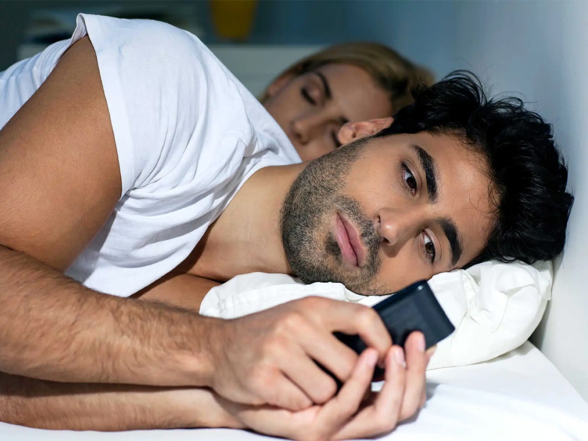 Man beside his partner in bed looking at his phone