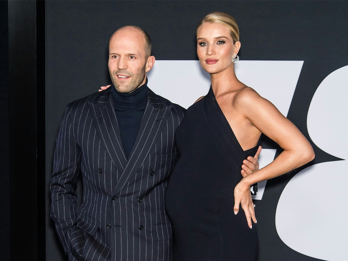 Jason Staham in a turtleneck underneath patterned suit with his pregnant partner in a black dress