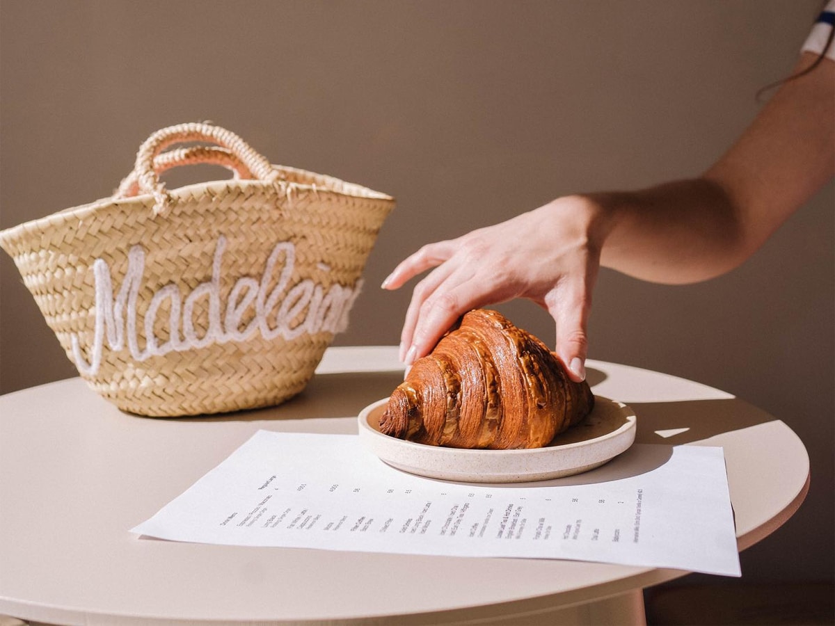 Hand holding a croissant set on a table beside a menu on a paper and a bag