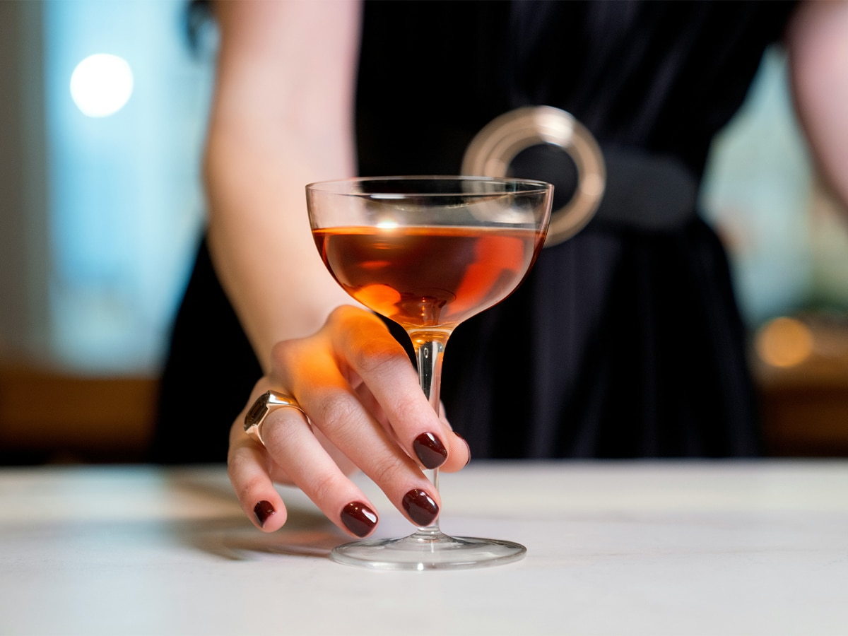 Woman holding a Manhattan cocktail in a coupe glass with a dark dress and red nails.
