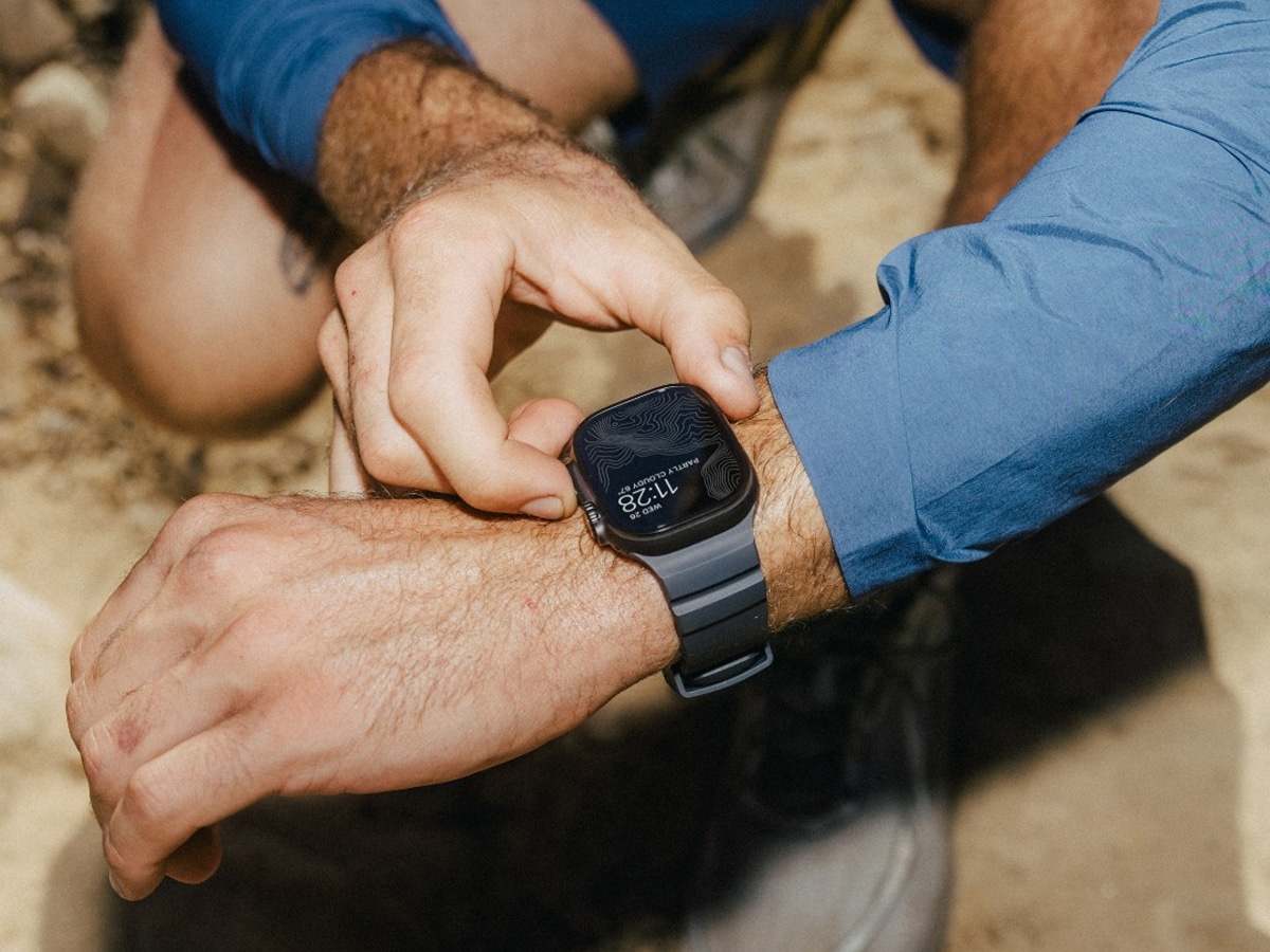 Person adjusting Apple Watch with black Nomad band, wearing a blue long-sleeve shirt outdoors.