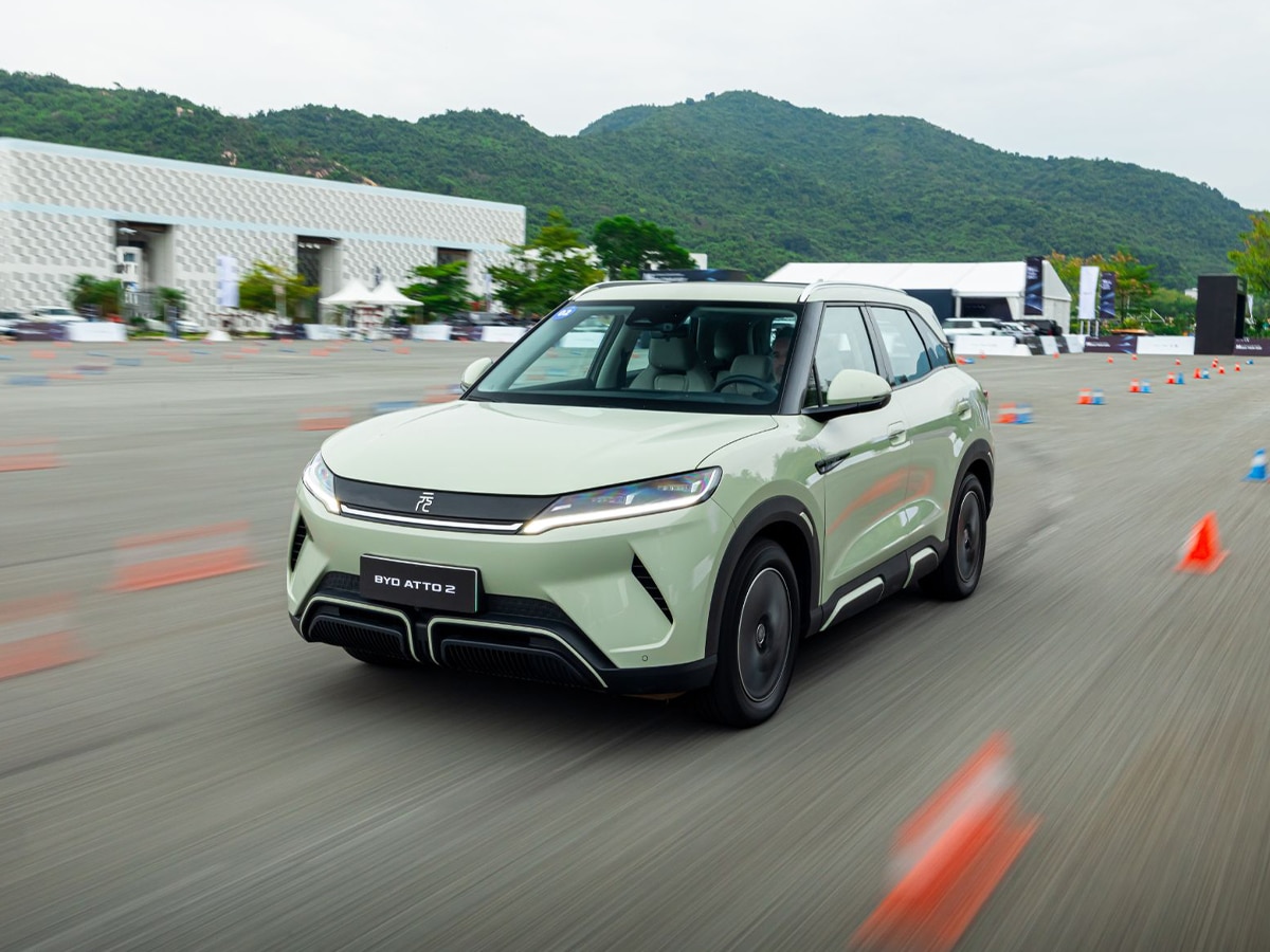BYD Atto 2 in light green driving on a test track with mountains and modern buildings in the background.