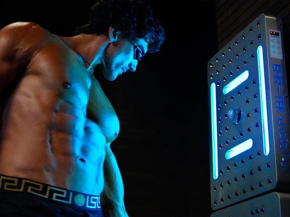 Muscular man in underwear and protective goggles standing close to a fully lit EMR-TEK device emitting bright blue light, in a dark indoor setting
