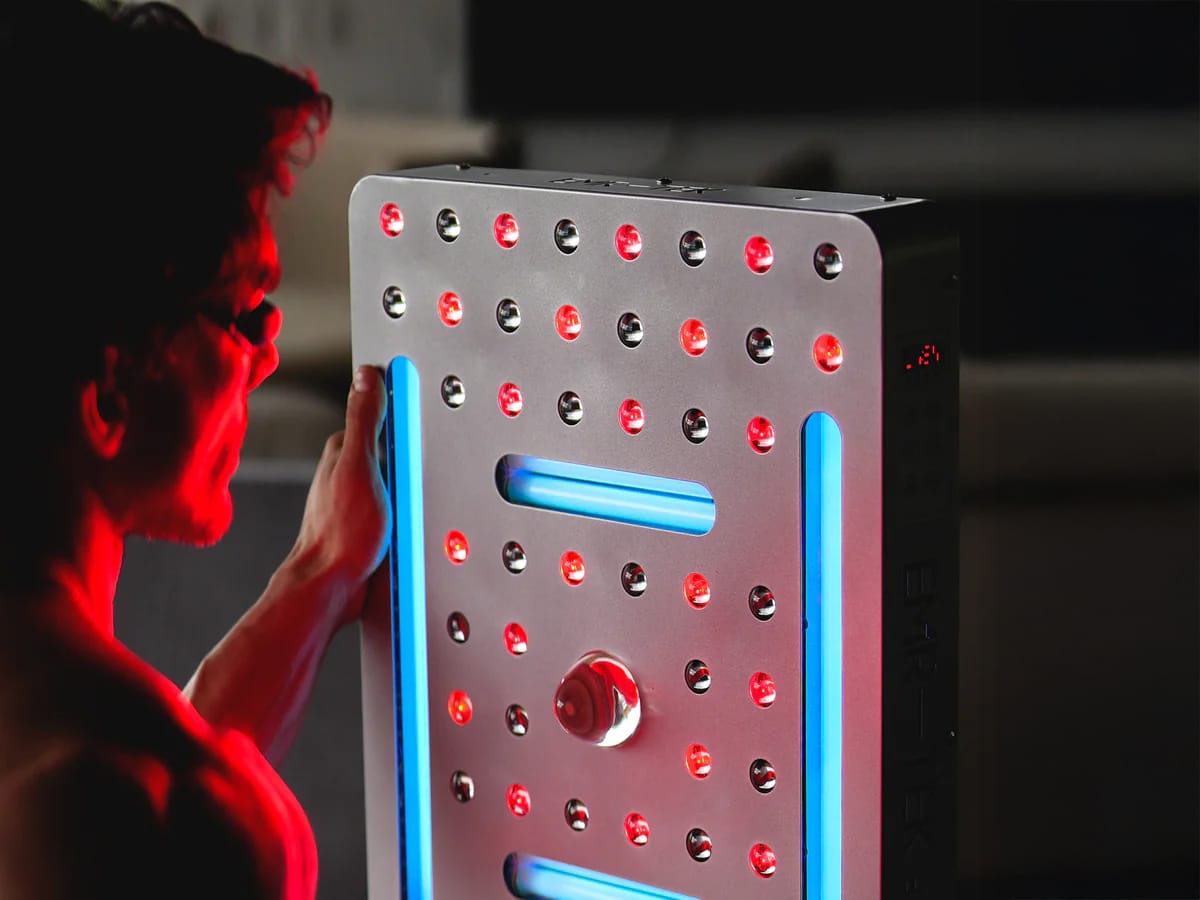 Close-up of a man adjusting or inspecting a red and blue LED light therapy device, wearing protective eyewear and surrounded by a red glow