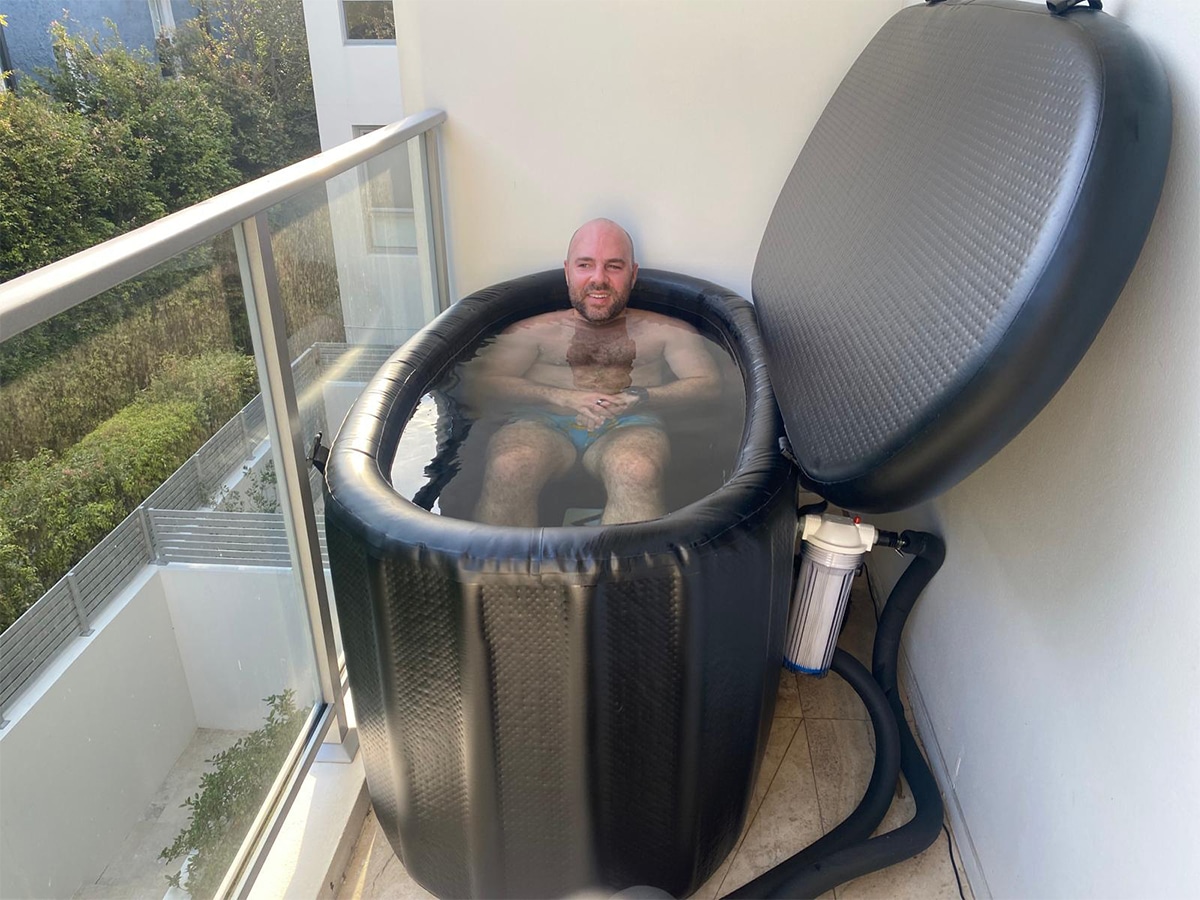 Man sitting in an inflatable ice bath on a balcony, partially submerged in water, and a water filtration system attached to the side
