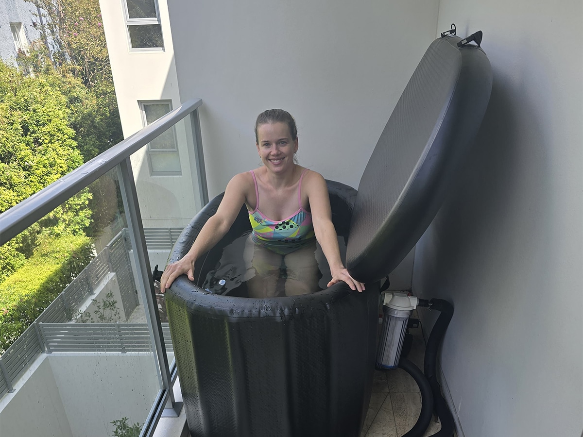 Woman in a colourful swimsuit smiling while sitting in an inflatable ice bath on a balcony, with trees and buildings visible in the background