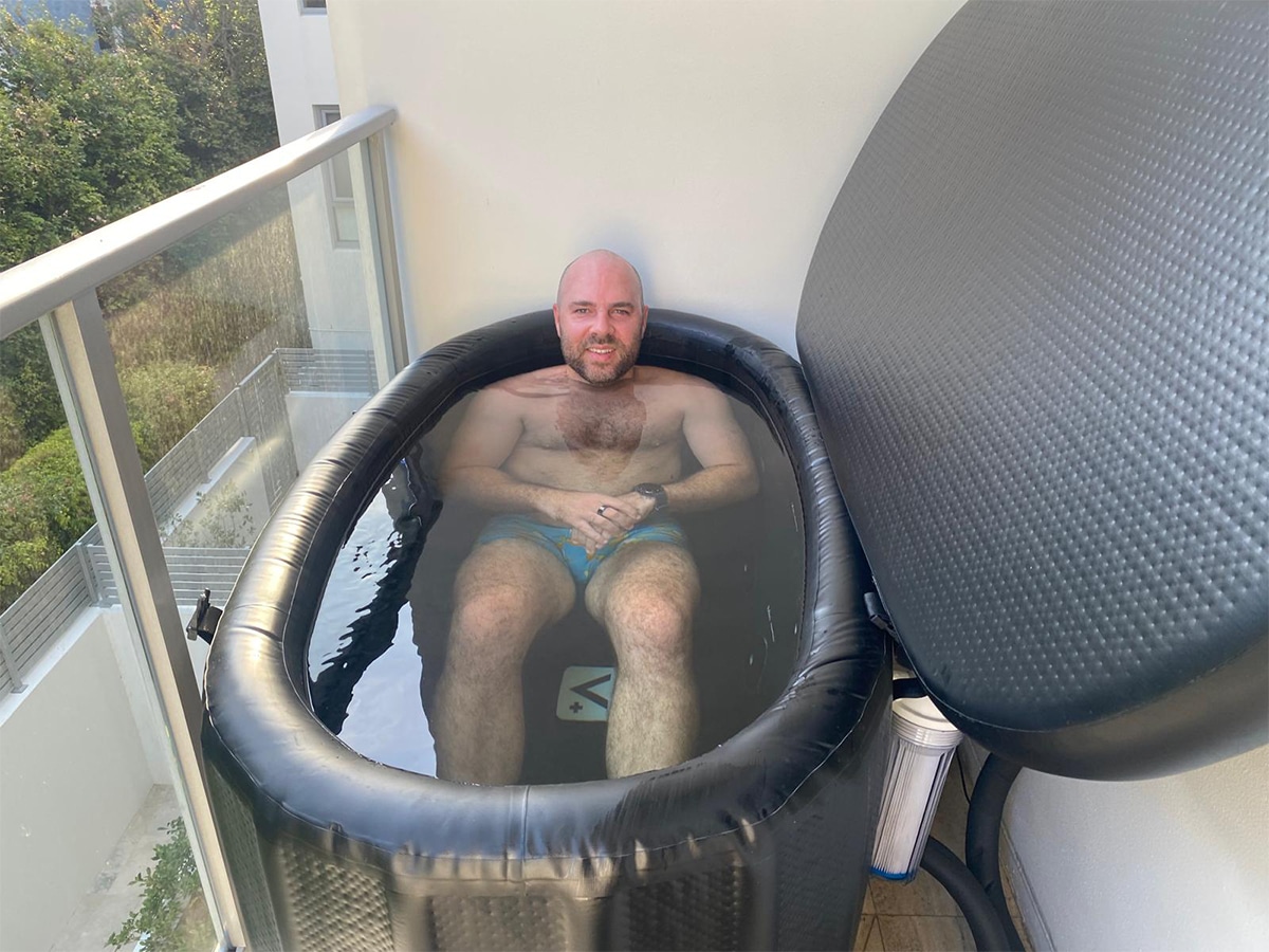 Man sitting in an inflatable ice bath on a balcony, partially submerged in water, and a water filtration system attached to the side