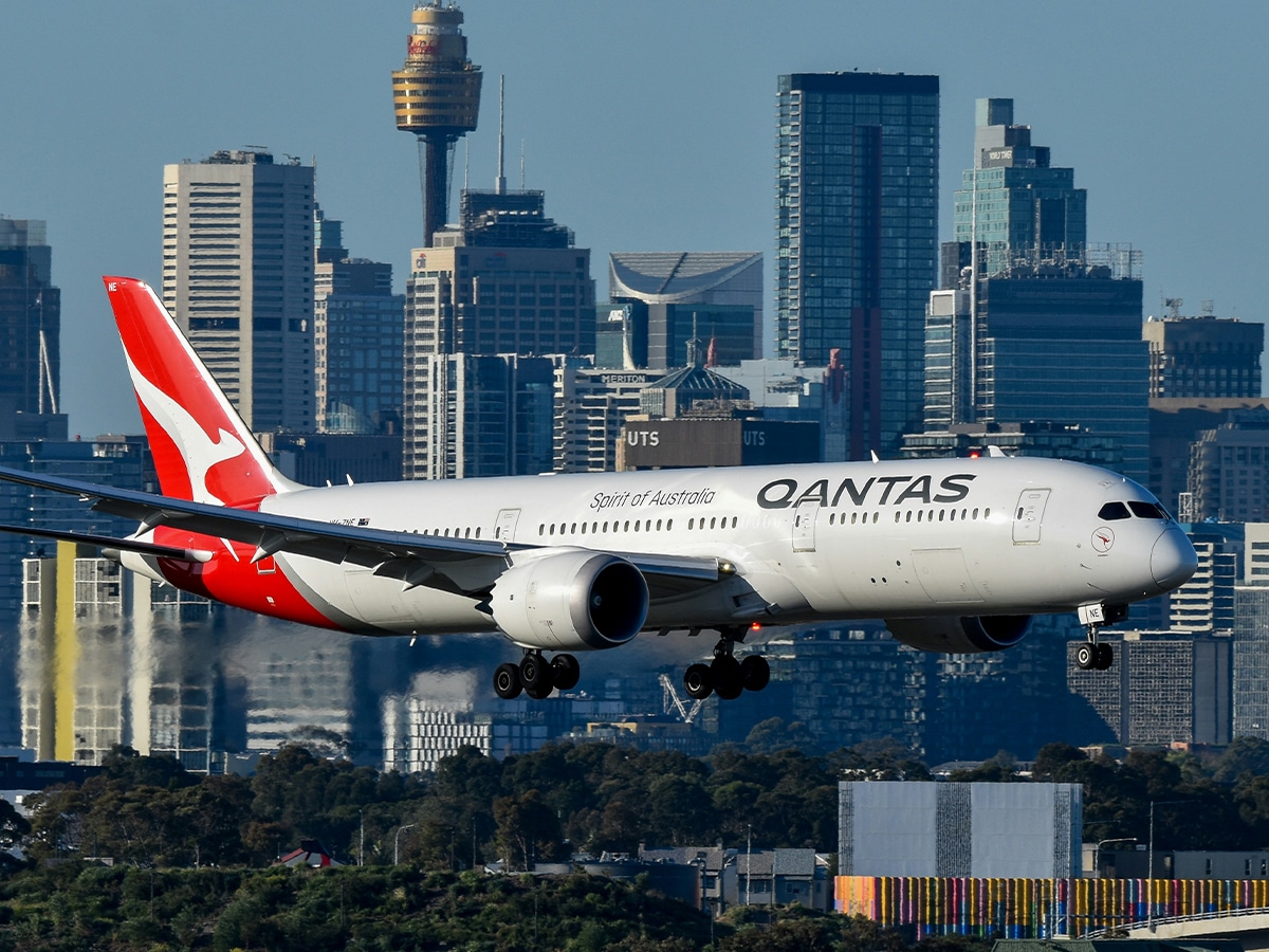 Qantas Boeing 787 Dreamliner landing with Sydney skyline in the background, featuring clear blue sky.