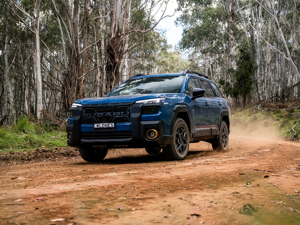 2026 Subaru Outback Wilderness in blue driving on a dirt road through a forest with trees in the background.
