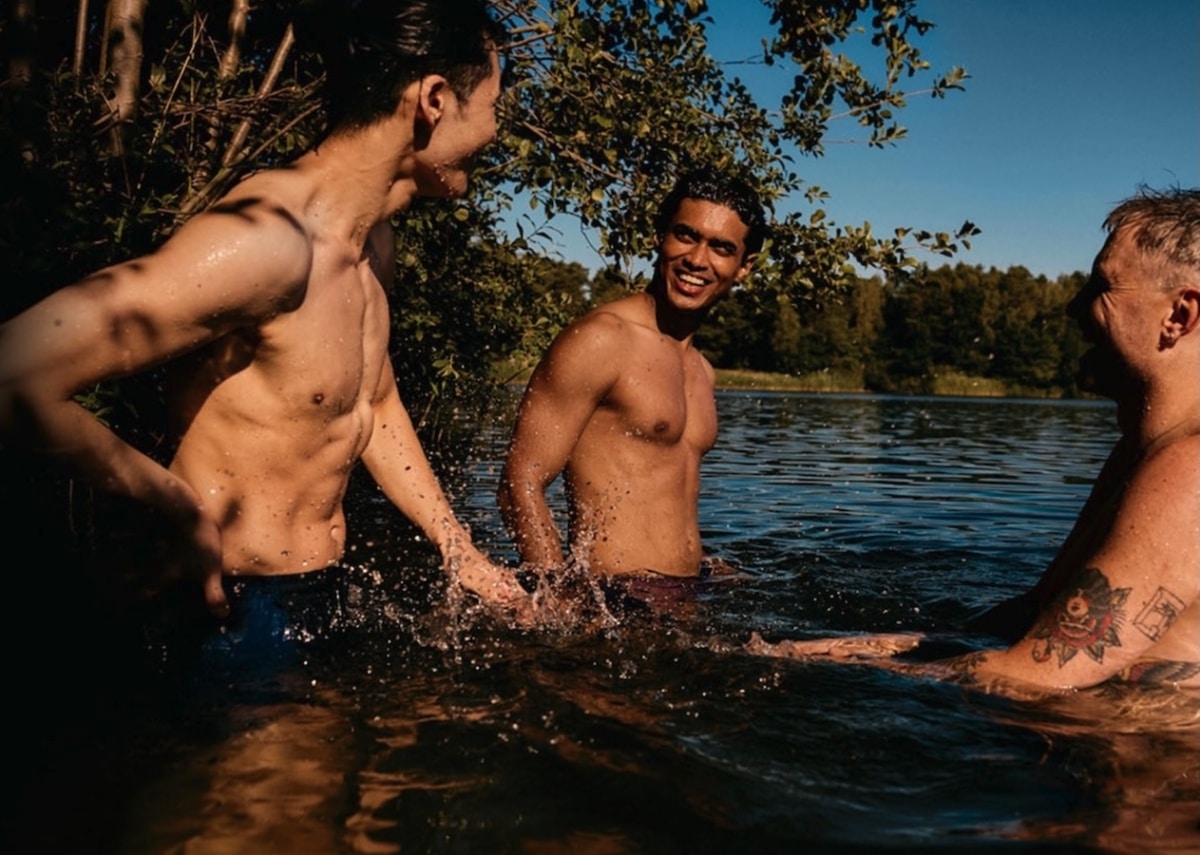 Three men smiling and standing in a sunlit lake, surrounded by trees, enjoying a swim together.