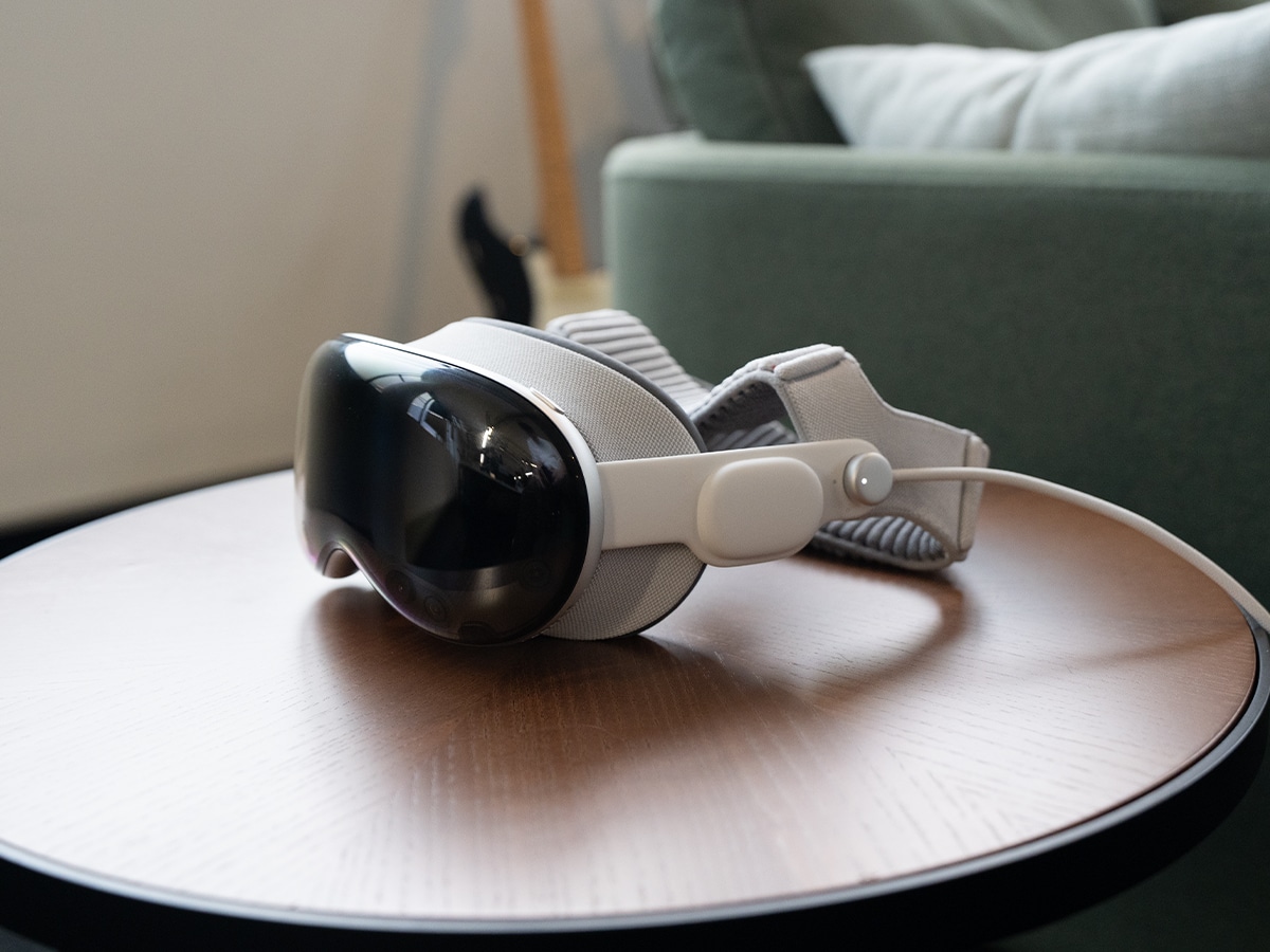 Apple Vision Pro headset on round wooden table, featuring sleek design and grey strap in a modern living room setting.